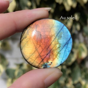 May include: A round, polished gemstone with a vibrant color gradient of yellow, orange, and blue. The stone has a network of dark lines across its surface, and the text "Au soleil" is visible.