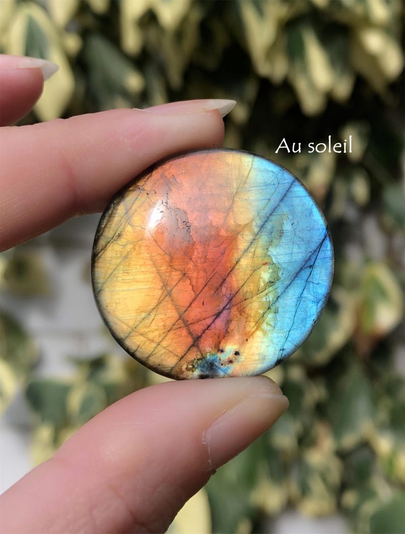 May include: A round, polished gemstone with a vibrant color gradient of yellow, orange, and blue. The stone has a network of dark lines across its surface, and the text "Au soleil" is visible.