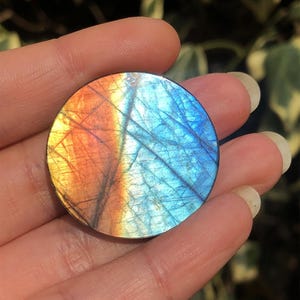 May include: A round, polished gemstone with a vibrant color gradient. The stone transitions from orange to blue, with intricate internal patterns. The gemstone is held in a hand, showcasing its size and detail.