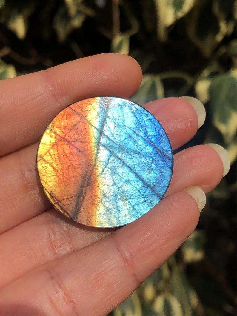 May include: A round, polished gemstone with a vibrant color gradient. The stone transitions from orange to blue, with intricate internal patterns. The gemstone is held in a hand, showcasing its size and detail.