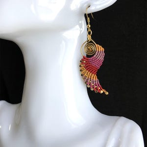 Pode incluir: Um par de brincos de ouro com um design em espiral rosa, laranja e amarelo. Os brincos s&atilde;o feitos com uma t&eacute;cnica de macram&eacute; e t&ecirc;m um gancho de ouro.