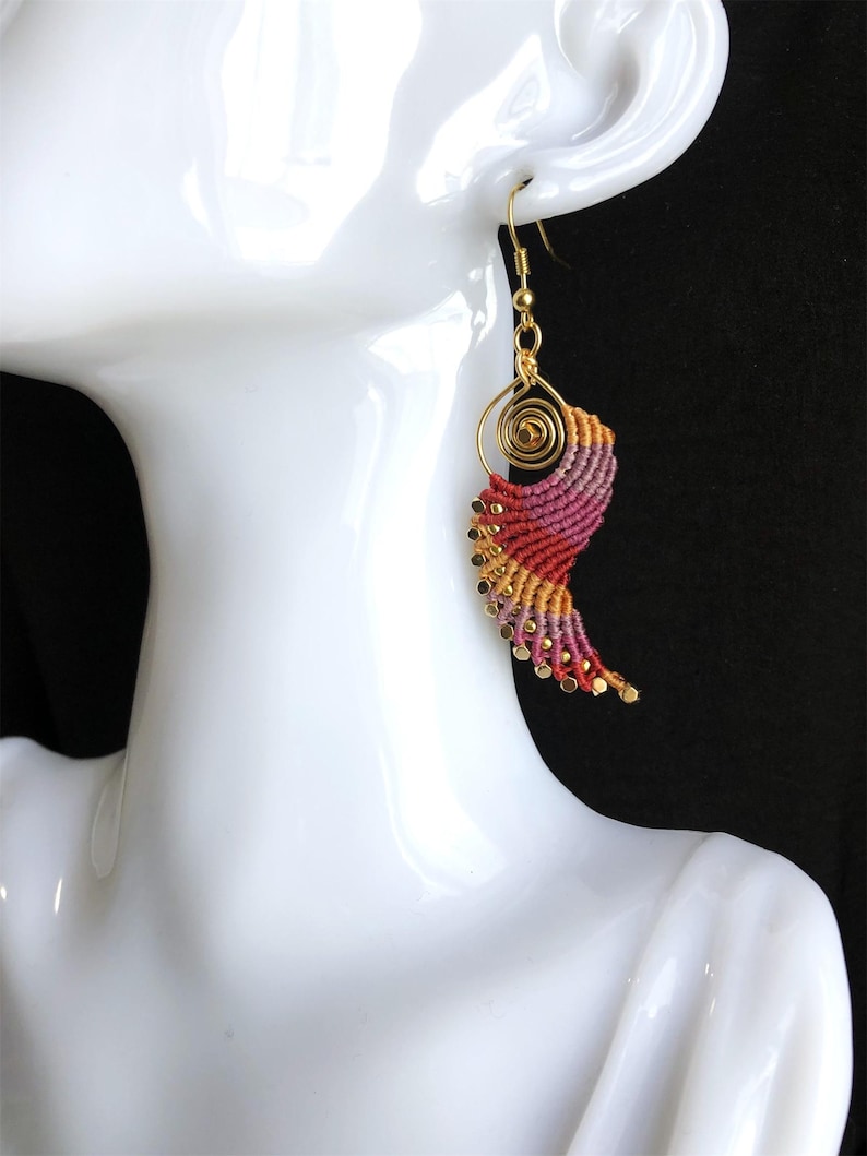 Pode incluir: Um par de brincos de ouro com um design em espiral rosa, laranja e amarelo. Os brincos s&atilde;o feitos com uma t&eacute;cnica de macram&eacute; e t&ecirc;m um gancho de ouro.