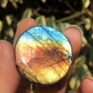 May include: A round, polished labradorite gemstone with a vibrant display of colors. The stone features a mix of blues, yellows, oranges, and browns, with a shimmering, iridescent effect. The gemstone is held between fingers, showcasing its natural beauty.
