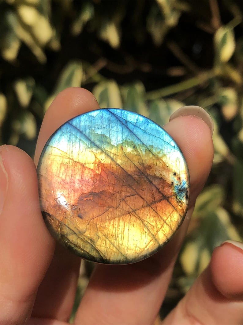 May include: A round, polished labradorite gemstone with a vibrant display of colors. The stone features a mix of blues, yellows, oranges, and browns, with a shimmering, iridescent effect. The gemstone is held between fingers, showcasing its natural beauty.