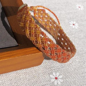 K&ouml;nnte beinhalten: Ein geflochtenes Armband mit einem komplexen Gittermuster aus orangefarbenen, gelben und beigen F&auml;den. Das Design enth&auml;lt einen dezenten metallischen Schimmer in den Fasern, wobei am Verschluss eine kleine, helle Perle zu sehen ist.