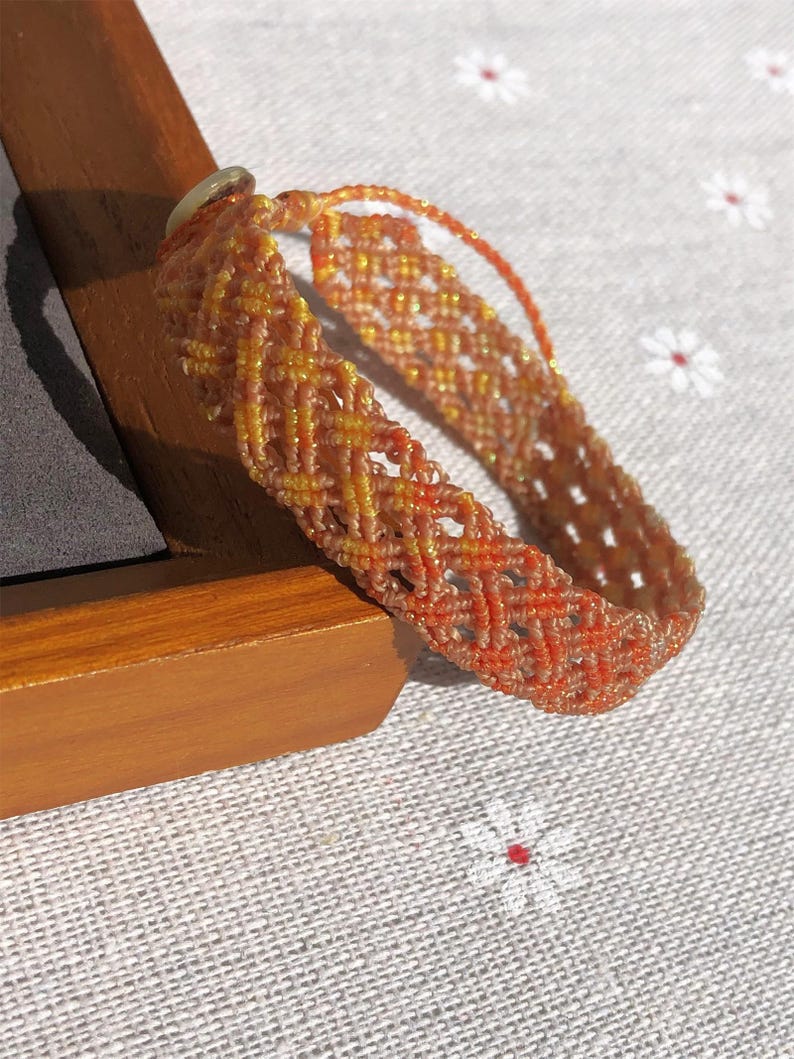 K&ouml;nnte beinhalten: Ein geflochtenes Armband mit einem komplexen Gittermuster aus orangefarbenen, gelben und beigen F&auml;den. Das Design enth&auml;lt einen dezenten metallischen Schimmer in den Fasern, wobei am Verschluss eine kleine, helle Perle zu sehen ist.
