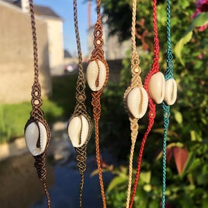 Può includere: Una collezione di collane in macramè, ognuna con un ciondolo a conchiglia bianca. Le collane variano di colore, tra cui marrone, beige, rosso e turchese. I nodi e i cordoncini in macramè creano un'estetica bohémien.