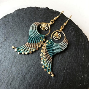 Pode incluir: Um par de brincos de macram&eacute; verde e marrom com contas douradas e argolas de arame. Os brincos est&atilde;o pendurados em uma superf&iacute;cie cinza escura.