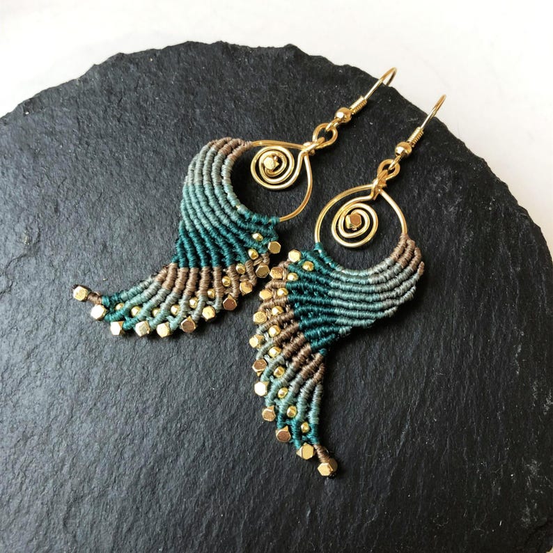 Pode incluir: Um par de brincos de macram&eacute; verde e marrom com contas douradas e argolas de arame. Os brincos est&atilde;o pendurados em uma superf&iacute;cie cinza escura.