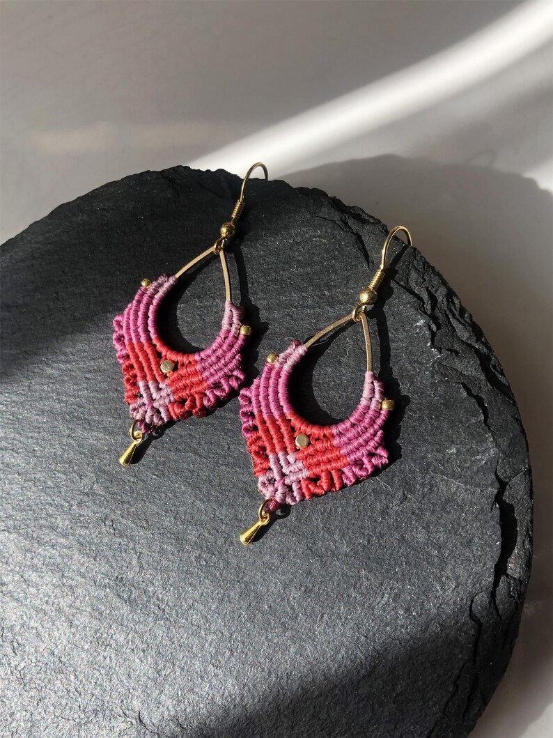 May include: A pair of handmade macrame earrings. The earrings feature a teardrop shape with a gradient of pink and red threads. Gold-colored metal accents and hooks complete the design, creating a bohemian style.