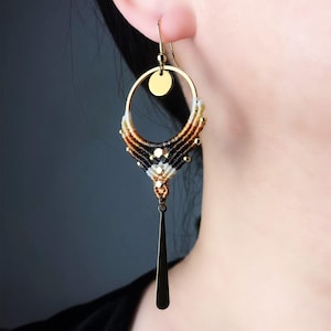 Peut inclure: Une boucle d'oreille pendante composée d'un anneau en métal doré avec une petite breloque circulaire en haut. La moitié inférieure de l'anneau est entourée d'un cordon macramé dégradé crème, beige, marron et noir, orné de perles dorées et d'une tige.