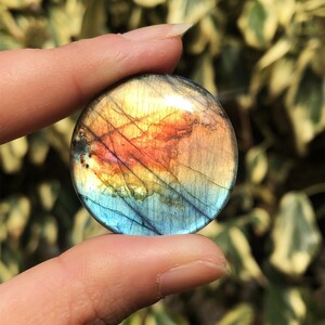 May include: A round, polished labradorite gemstone with a vibrant display of colors. The stone features a mix of blue, orange, and yellow hues, with subtle lines and patterns. The gemstone is held between two fingers.