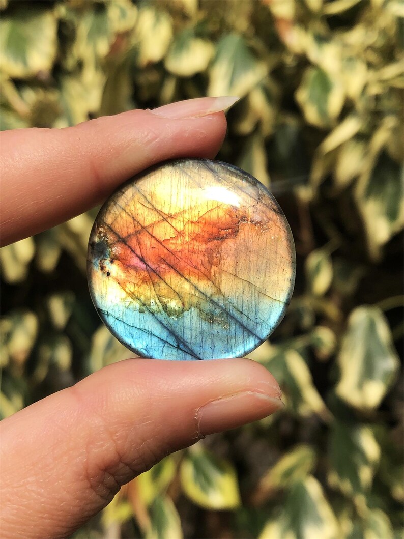 May include: A round, polished labradorite gemstone with a vibrant display of colors. The stone features a mix of blue, orange, and yellow hues, with subtle lines and patterns. The gemstone is held between two fingers.