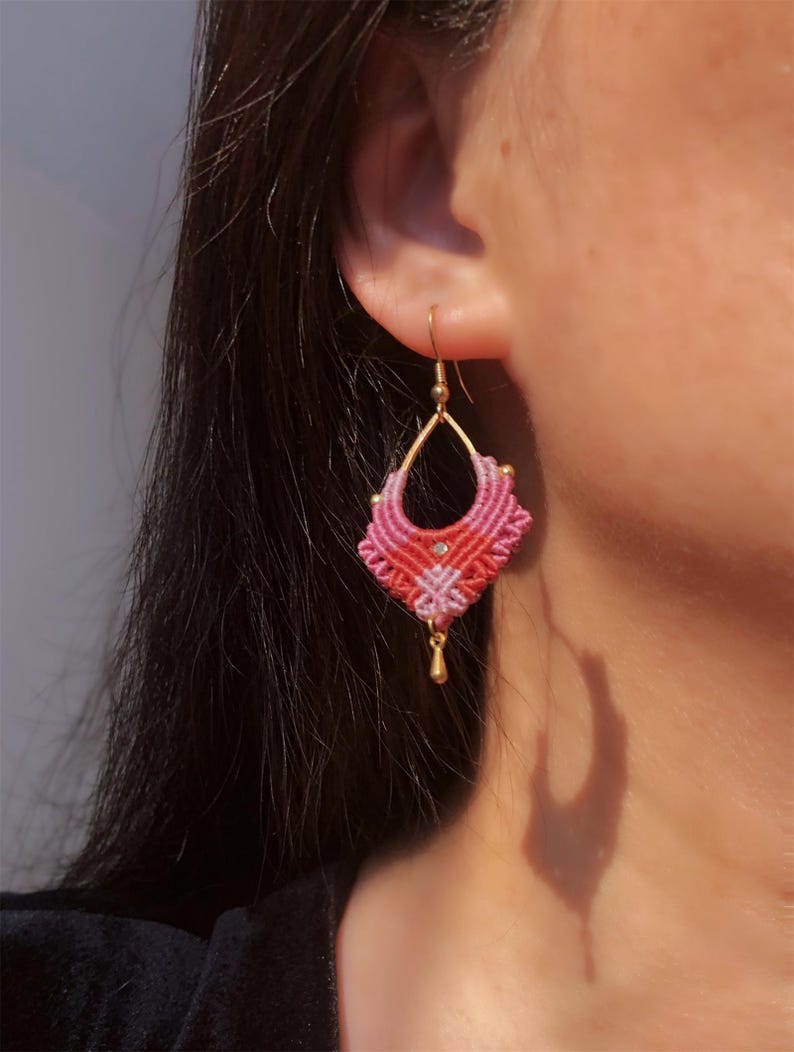 May include: A close-up of a pink and red macrame earring with a gold-colored hook and small gold bead. The earring has a geometric design and is hanging from an ear.
