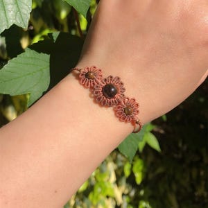 Peut inclure: Bracelet en macramé fait à la main avec trois motifs en forme de fleurs. Le bracelet est composé d'un cordon marron et comporte une pierre centrale brun foncé, avec des pierres brun clair dans les autres fleurs. Le bracelet est porté au poignet.