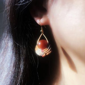 May include: A single earring with a gold-colored teardrop frame holding a round, reddish-orange stone. The stone is surrounded by a macrame design in cream, tan, and dark brown. The earring is suspended from a gold-colored hook.