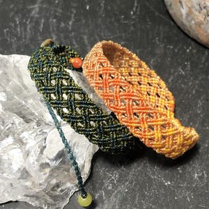 K&ouml;nnte beinhalten: Zwei Makramee-Armb&auml;nder mit Gittermuster. Ein Armband ist dunkelgr&uuml;n mit einer kleinen roten Perle und einer herabh&auml;ngenden hellgr&uuml;nen Perle. Das zweite Armband zeigt einen Farbverlauf in Orange- und Gelbt&ouml;nen. Beide sind aus d&uuml;nner Kordel gefertigt.