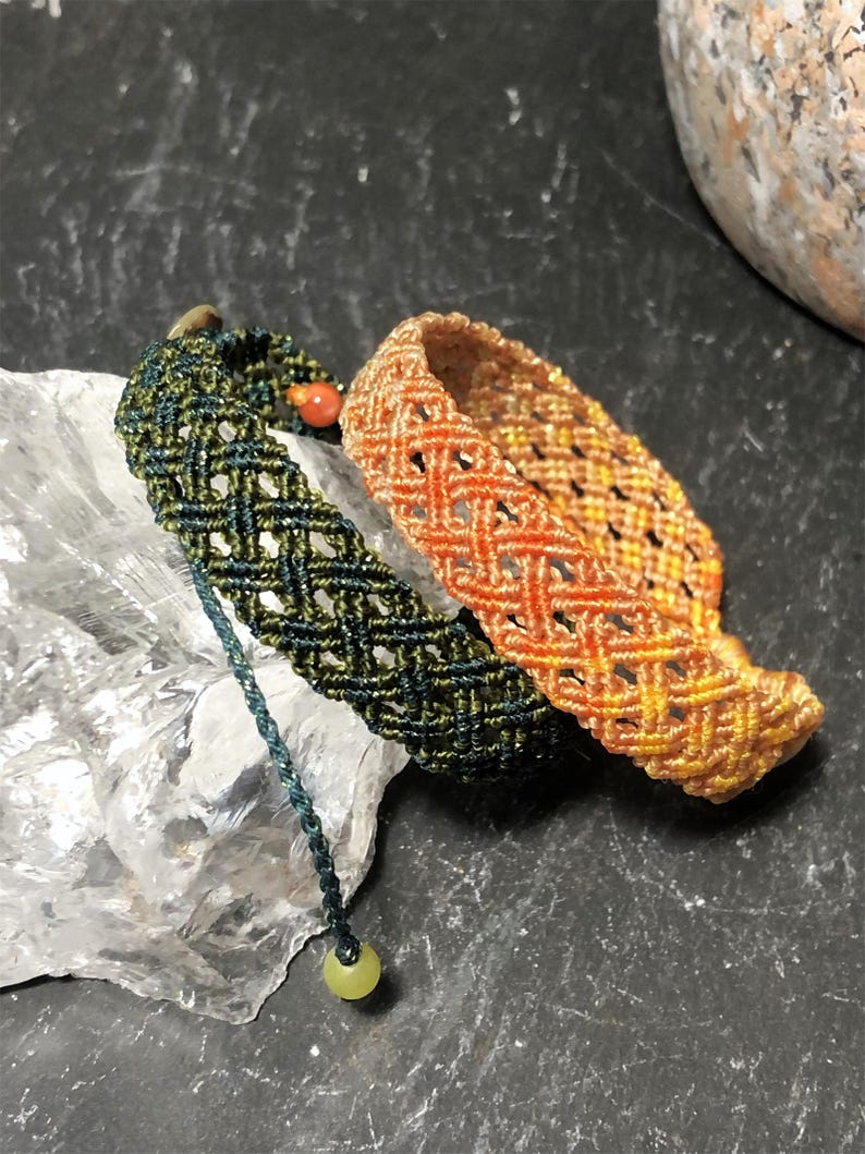 K&ouml;nnte beinhalten: Zwei Makramee-Armb&auml;nder mit Gittermuster. Ein Armband ist dunkelgr&uuml;n mit einer kleinen roten Perle und einer herabh&auml;ngenden hellgr&uuml;nen Perle. Das zweite Armband zeigt einen Farbverlauf in Orange- und Gelbt&ouml;nen. Beide sind aus d&uuml;nner Kordel gefertigt.
