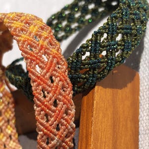 K&ouml;nnte beinhalten: Zwei geflochtene Armb&auml;nder mit komplexen Makramee-Knotenmustern. Ein Armband zeigt einen Farbverlauf in Pfirsich- und Oranget&ouml;nen mit dezentem metallischem Schimmer, w&auml;hrend das andere in tiefem Waldgr&uuml;n mit durchgehendem Glitzerfaden gehalten ist.