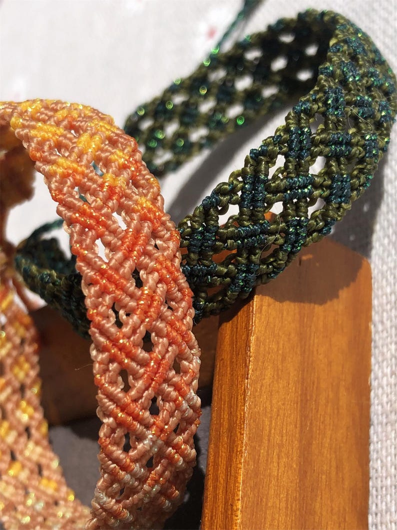 K&ouml;nnte beinhalten: Zwei geflochtene Armb&auml;nder mit komplexen Makramee-Knotenmustern. Ein Armband zeigt einen Farbverlauf in Pfirsich- und Oranget&ouml;nen mit dezentem metallischem Schimmer, w&auml;hrend das andere in tiefem Waldgr&uuml;n mit durchgehendem Glitzerfaden gehalten ist.