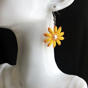 May include: Yellow daisy flower earrings with a white pearl center and orange accents. The earrings are hanging from silver hooks.