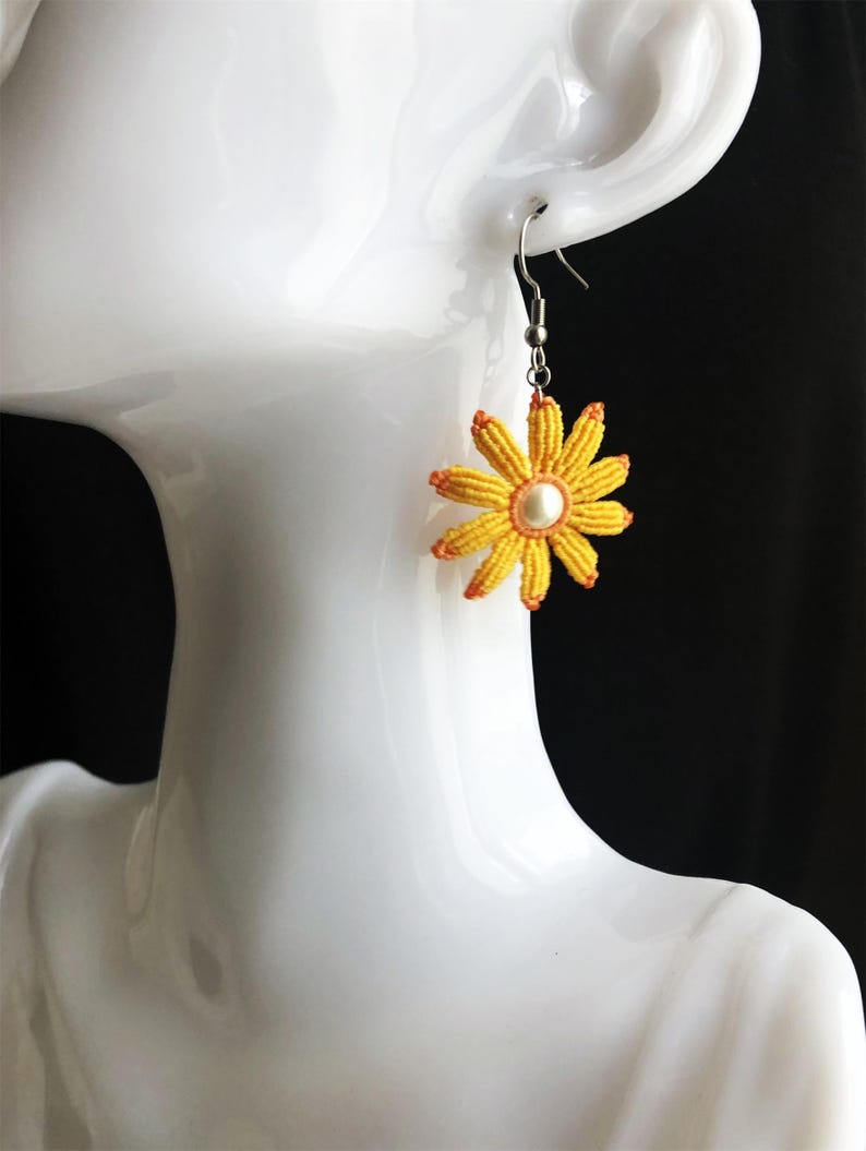 May include: Yellow daisy flower earrings with a white pearl center and orange accents. The earrings are hanging from silver hooks.