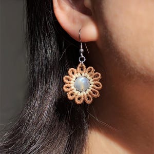 May include: A close-up of a handmade earring. The earring features a flower design with a light brown macrame outer layer and a light blue gemstone centre. The earring is attached to a silver hook.
