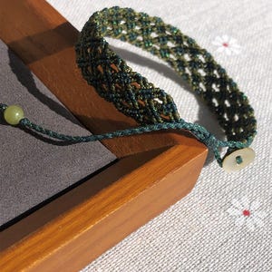 K&ouml;nnte beinhalten: Ein geflochtenes Armband mit einem Gittermuster aus dunkelgr&uuml;nen und olivfarbenen F&auml;den. Das Armband verf&uuml;gt &uuml;ber eine hellgr&uuml;ne Perle an einem Ende der Schnur und einen kleinen, runden, cremewei&szlig;en Knopf am anderen Ende zum Verschlie&szlig;en.