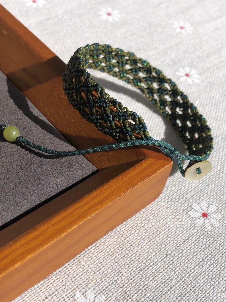 K&ouml;nnte beinhalten: Ein geflochtenes Armband mit einem Gittermuster aus dunkelgr&uuml;nen und olivfarbenen F&auml;den. Das Armband verf&uuml;gt &uuml;ber eine hellgr&uuml;ne Perle an einem Ende der Schnur und einen kleinen, runden, cremewei&szlig;en Knopf am anderen Ende zum Verschlie&szlig;en.