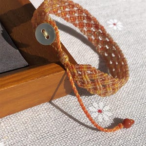K&ouml;nnte beinhalten: Ein geflochtenes Armband mit einem Gittermuster in Orange- und Gelbt&ouml;nen. Das Armband wird mit einem einzelnen wei&szlig;en, runden Knopf verschlossen und verf&uuml;gt &uuml;ber eine d&uuml;nne, geflochtene Kordelverl&auml;ngerung, die in einer kleinen, glatten roten Perle endet.