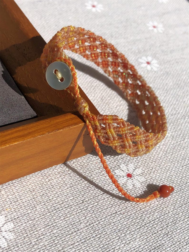 K&ouml;nnte beinhalten: Ein geflochtenes Armband mit einem Gittermuster in Orange- und Gelbt&ouml;nen. Das Armband wird mit einem einzelnen wei&szlig;en, runden Knopf verschlossen und verf&uuml;gt &uuml;ber eine d&uuml;nne, geflochtene Kordelverl&auml;ngerung, die in einer kleinen, glatten roten Perle endet.