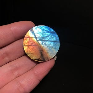 May include: A round, polished labradorite stone displays a vibrant array of colors. The stone's surface showcases a blend of blues, oranges, and yellows, with intricate patterns. The gemstone is held in a hand against a black background.