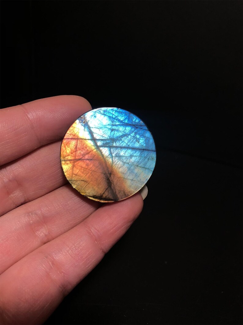 May include: A round, polished labradorite stone displays a vibrant array of colors. The stone's surface showcases a blend of blues, oranges, and yellows, with intricate patterns. The gemstone is held in a hand against a black background.