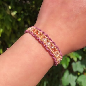 May include: A macrame bracelet with a woven pattern, alternating light pink and beige cord. It features small, round, translucent beads in yellow and peach. The bracelet is worn on a wrist. The bracelet is approximately 1.5 cm wide.