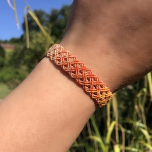 K&ouml;nnte beinhalten: Ein gewebtes Makramee-Armband mit Rautenmuster in einem Farbverlauf von hellem Creme bis hin zu leuchtendem Orange und Gelb. Das Armband besteht aus d&uuml;nnem, geknotetem Garn, das ein strukturiertes, gitterartiges Design erzeugt.