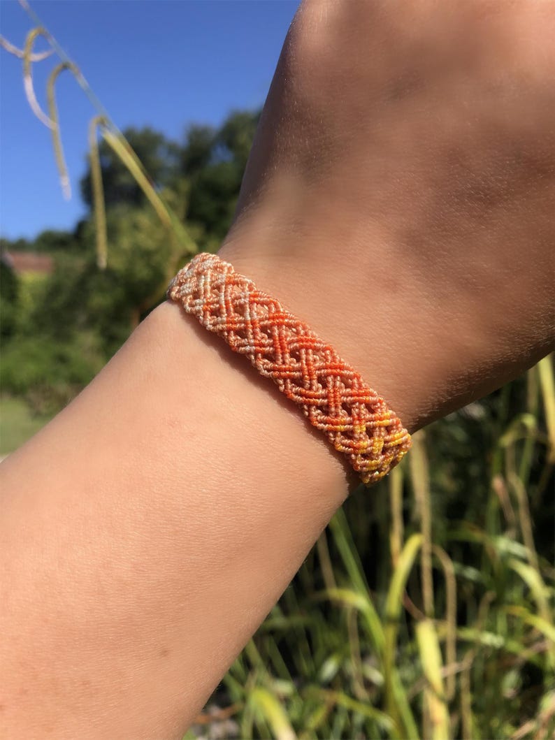 K&ouml;nnte beinhalten: Ein gewebtes Makramee-Armband mit Rautenmuster in einem Farbverlauf von hellem Creme bis hin zu leuchtendem Orange und Gelb. Das Armband besteht aus d&uuml;nnem, geknotetem Garn, das ein strukturiertes, gitterartiges Design erzeugt.