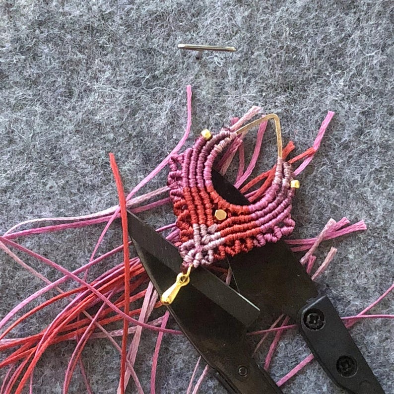 May include: A handmade macrame earring in shades of red and pink, with gold-colored accents. The earring is positioned on top of black scissors, surrounded by strands of pink and red thread, against a gray felt background.