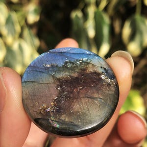 May include: A round, polished labradorite gemstone with a mix of blue, purple, and gold colors. The stone has a smooth surface and displays iridescent flashes. The gemstone is held in a hand against a blurred green background.