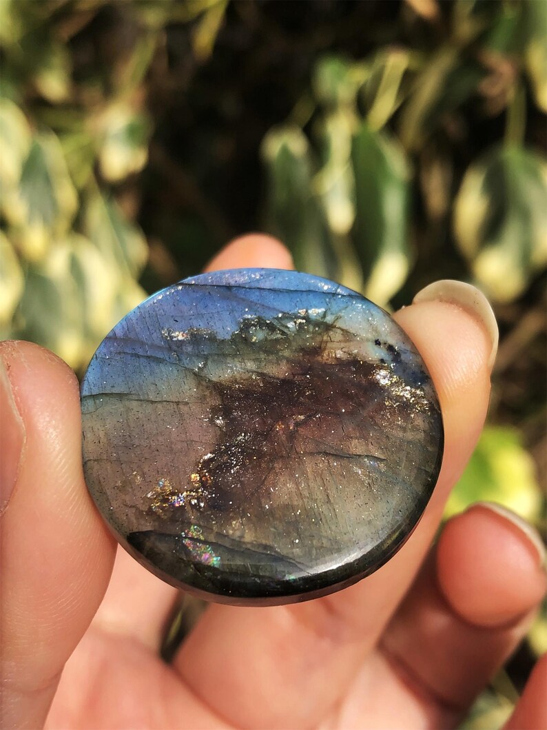 May include: A round, polished labradorite gemstone with a mix of blue, purple, and gold colors. The stone has a smooth surface and displays iridescent flashes. The gemstone is held in a hand against a blurred green background.