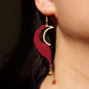 Peut inclure: Une boucle d'oreille unique avec un croissant de lune doré et un motif en forme d'aile en macramé rouge foncé. Une petite perle dorée est suspendue en bas. La boucle d'oreille est suspendue à un crochet doré.