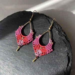 Könnte beinhalten: Ein Paar handgefertigte Makramee-Ohrringe. Die Ohrringe haben eine Tropfenform mit einem goldfarbenen Metallrahmen und einem gewebten Muster in Rosa- und Rottönen. Kleine goldene Perlen akzentuieren das Design.