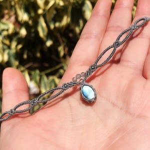 May include: A grey macramé bracelet with a light blue oval gemstone pendant. The bracelet has a delicate woven design with small, dark grey beads. The gemstone is a focal point, adding a touch of elegance to the accessory.