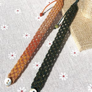 K&ouml;nnte beinhalten: Zwei Makramee-Armb&auml;nder mit geflochtenem Gittermuster. Ein Armband ist orange und gelb, das andere dunkelgr&uuml;n. Jedes Armband hat einen wei&szlig;en, runden Knopfverschluss an einem Ende und eine kleine Zierperle an der verstellbaren Kordel.