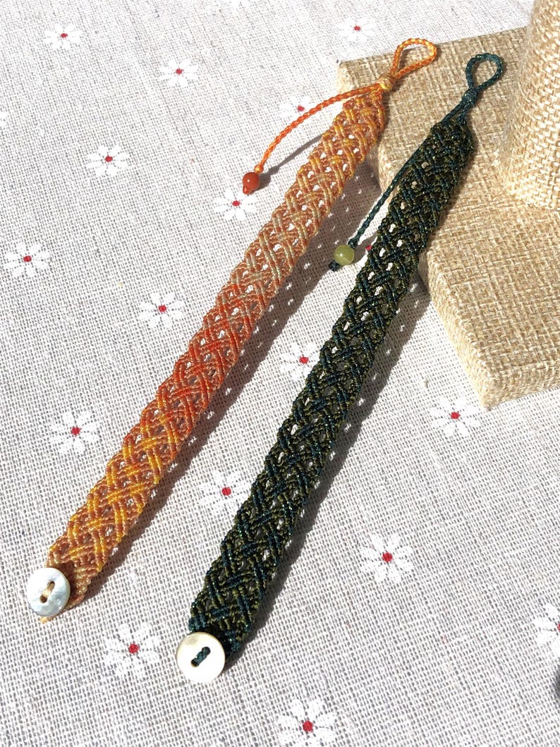 K&ouml;nnte beinhalten: Zwei Makramee-Armb&auml;nder mit geflochtenem Gittermuster. Ein Armband ist orange und gelb, das andere dunkelgr&uuml;n. Jedes Armband hat einen wei&szlig;en, runden Knopfverschluss an einem Ende und eine kleine Zierperle an der verstellbaren Kordel.