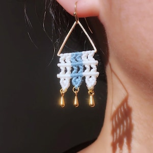 May include: A close-up of a macrame earring. The earring features a gold triangle top with white and light blue woven details. Three gold teardrop charms dangle from the bottom. The earring is worn on a person's ear.