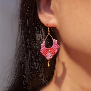 May include: A close-up of a pink and red macrame earring with a gold-colored hook and accents. The earring has a geometric design with a teardrop shape and a small gold bead at the bottom.