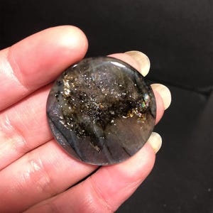 May include: A round, polished gemstone with a dark, iridescent surface. The stone features a mix of black, gray, and gold flecks, with hints of rainbow iridescence. The gemstone is held in a hand against a dark background.