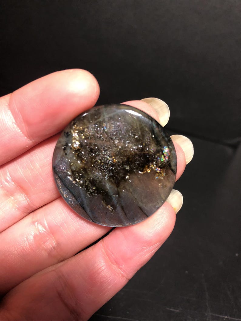 May include: A round, polished gemstone with a dark, iridescent surface. The stone features a mix of black, gray, and gold flecks, with hints of rainbow iridescence. The gemstone is held in a hand against a dark background.