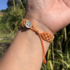 K&ouml;nnte beinhalten: Ein gewebtes Makramee-Armband mit einem lebendigen orange-gelben geometrischen Muster. Das Armband wird mit einem kleinen, runden, wei&szlig;en Perlmuttknopf verschlossen und hat eine d&uuml;nne orangefarbene Kordel, die in einer einzelnen, kleinen, roten Perle endet.