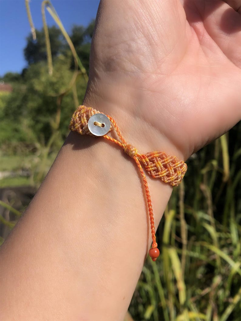 K&ouml;nnte beinhalten: Ein gewebtes Makramee-Armband mit einem lebendigen orange-gelben geometrischen Muster. Das Armband wird mit einem kleinen, runden, wei&szlig;en Perlmuttknopf verschlossen und hat eine d&uuml;nne orangefarbene Kordel, die in einer einzelnen, kleinen, roten Perle endet.